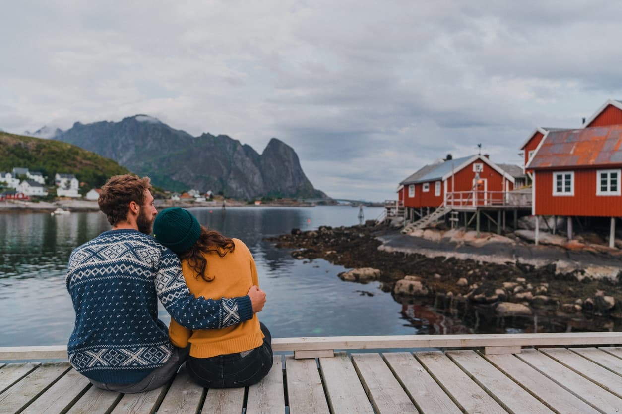 Et par sitter tett sammen på en brygge og ser utover vannet med røde rorbuer og fjell i bakgrunnen. Bildet illustrerer trygghet og fellesskap i et samboerforhold og kan brukes i innhold om arv for samboere uten felles barn.