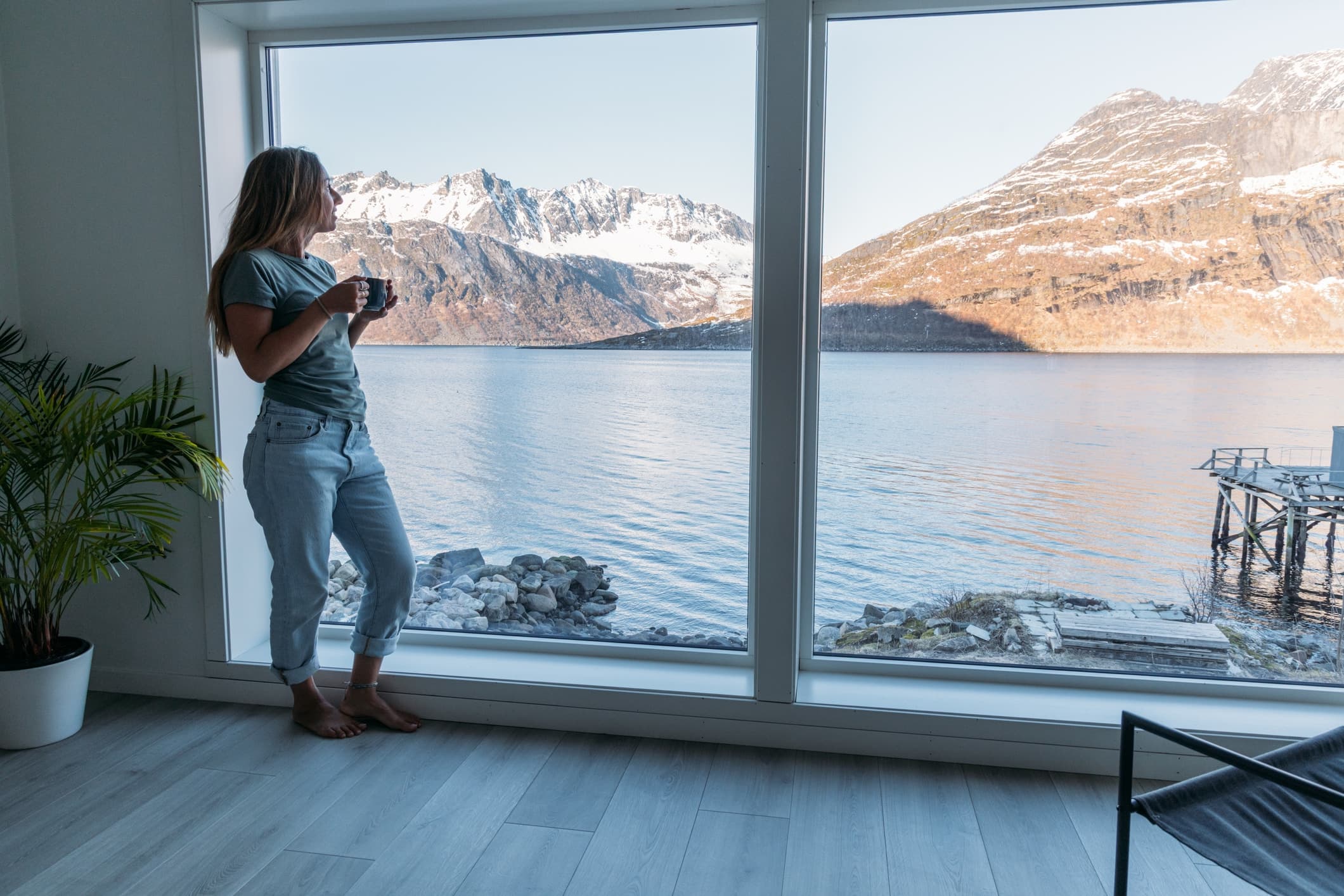 Kvinne står innendørs ved et stort vindu og ser utover fjord og fjell mens hun holder en kaffekopp. Bildet gir en rolig og ettertenksom stemning som kan knyttes til økonomiske og personlige valg i livet. Det fungerer som en visuell metafor for arv fra foreldre som særeie og langsiktige beslutninger knyttet til eiendom og verdier.